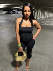 Full-length portrait of a Black woman standing against an off-white backdrop, wearing a black halter-neck crop top with a square front and matching high-waisted capri-length leggings that flare slightly at the hem. Her natural curls frame her face, she wears black strappy heels, and the clean background highlights the bold silhouette of the set.