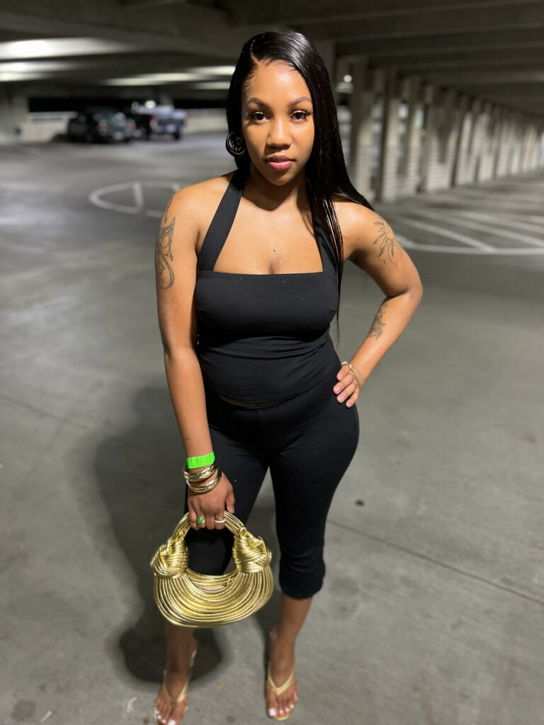 Full-length portrait of a Black woman standing against an off-white backdrop, wearing a black halter-neck crop top with a square front and matching high-waisted capri-length leggings that flare slightly at the hem. Her natural curls frame her face, she wears black strappy heels, and the clean background highlights the bold silhouette of the set.
