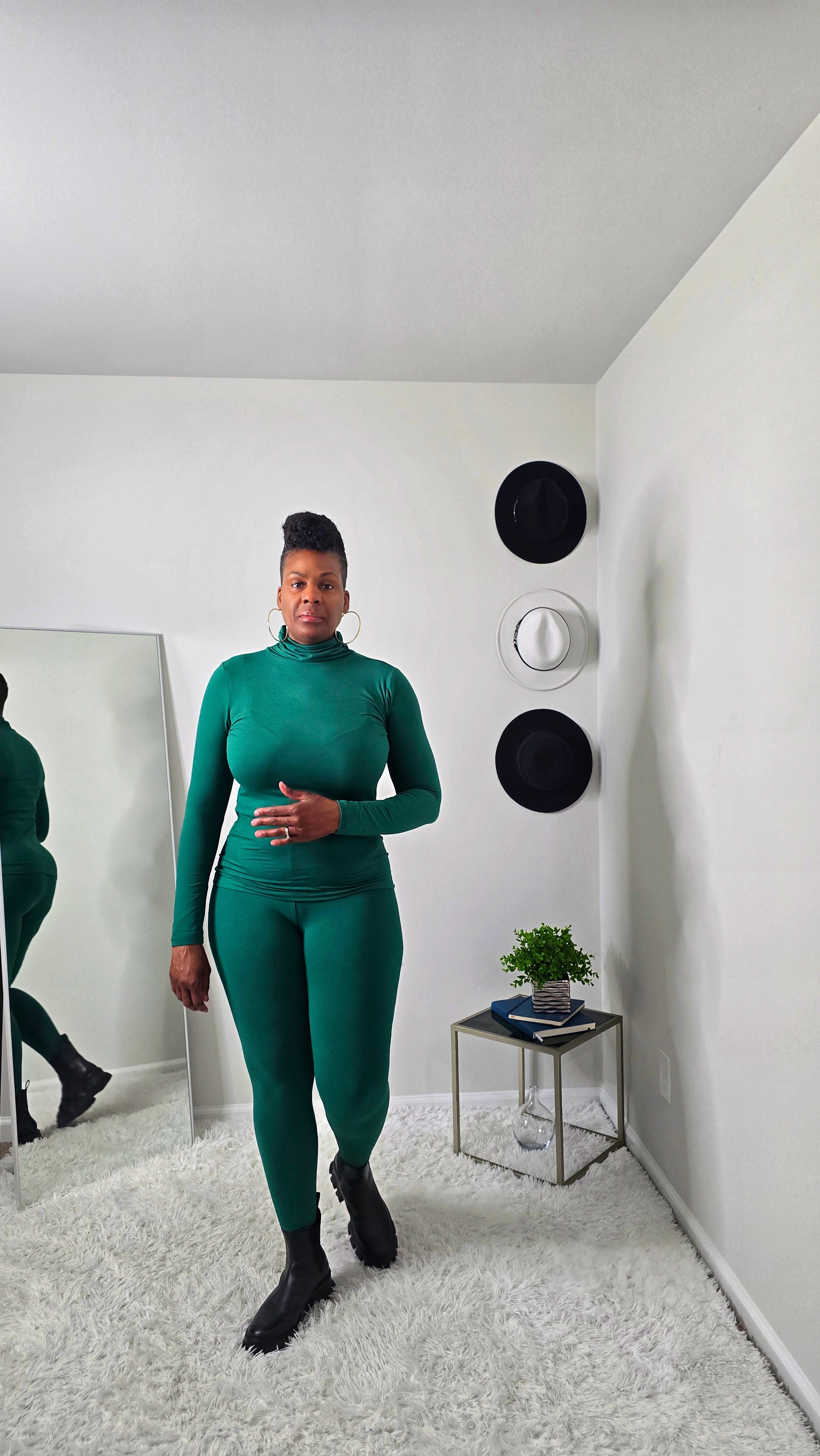 Person wearing a green outfit standing in a room with white walls and a mirror.