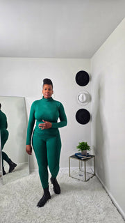 Person wearing a green outfit standing in a room with white walls and a mirror.