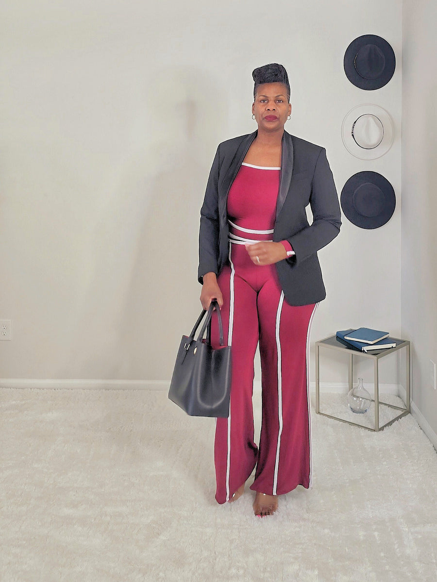 Woman in a black blazer and 2pc burgundy pant set standing in a room with a white wall and small table.