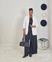 Woman in a white cardigan and black pants holding a black handbag in a minimalistic room.